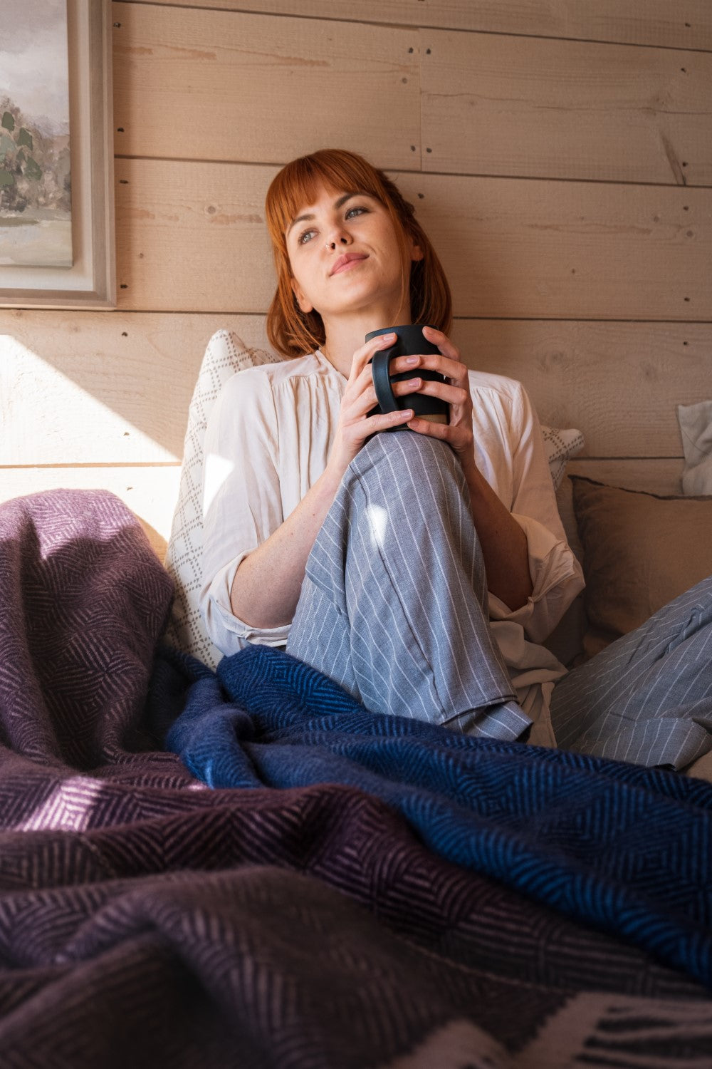 A woman sitting against a wooden wall panels with wool blankets draped on her left. 