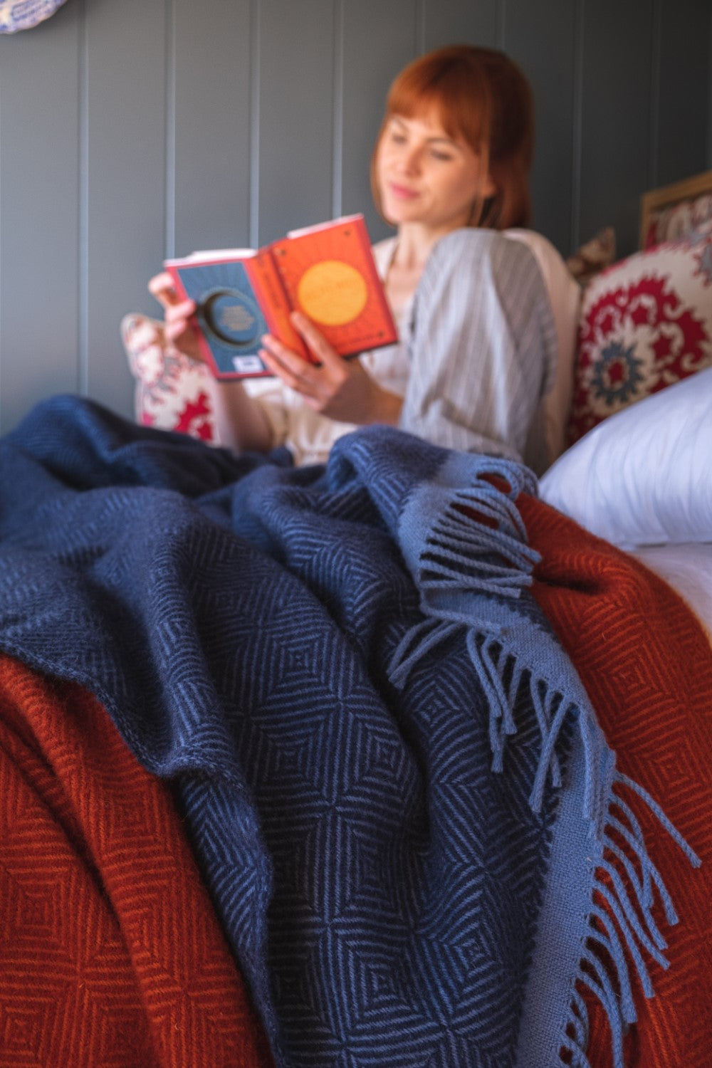 Blue and red wildweave wool blankets draped over a bed. A woman sitting cross legged is reading a book in the background