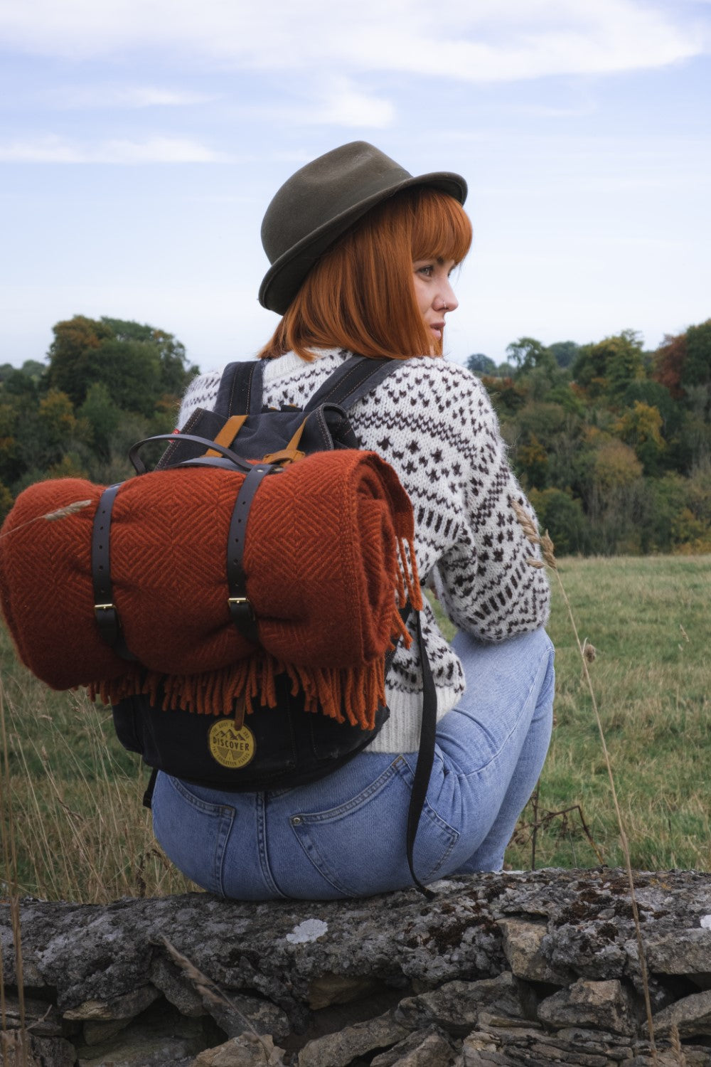 A woman sitting on a log with a rolled up red wildweave wool throw strapped to her backpack