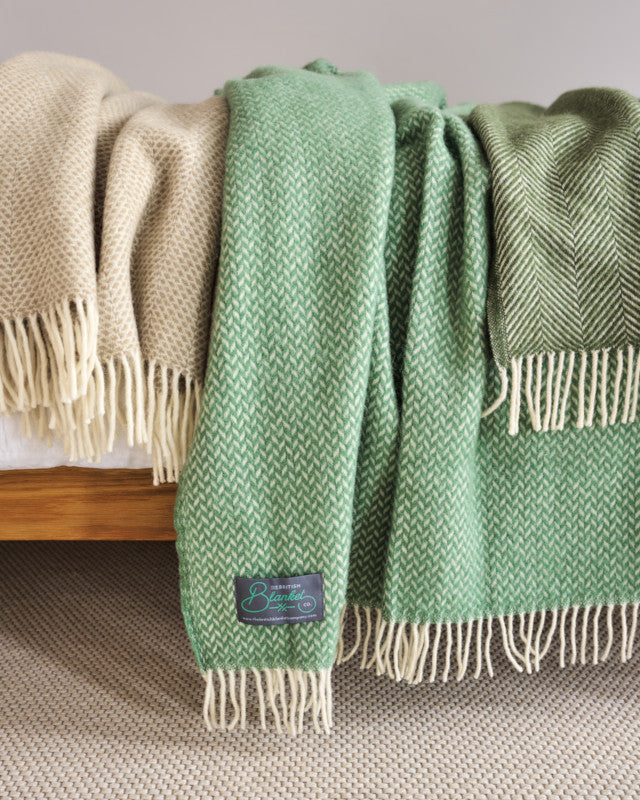 selection of natural beige and green wool throw blankets draped over a bed by The British Blanket Company online shop