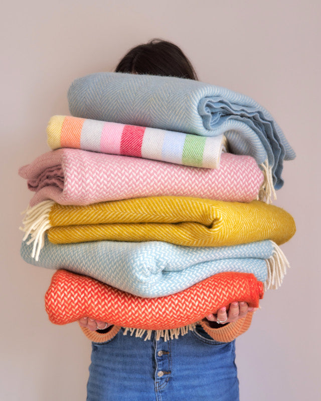 woman holding a very tall pile of colourful wool throw blankets by The British Blanket Company online shop