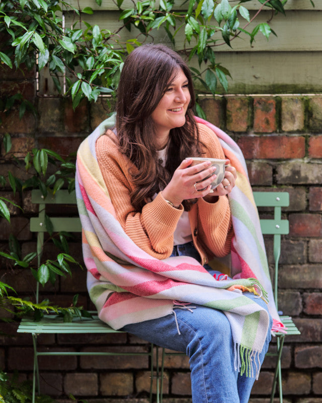 happy smiling woman sitting in a garden holding a cup of tea and wrapped in a rainbow stripe merino wool throw blanket by The British Blanket Company online shop