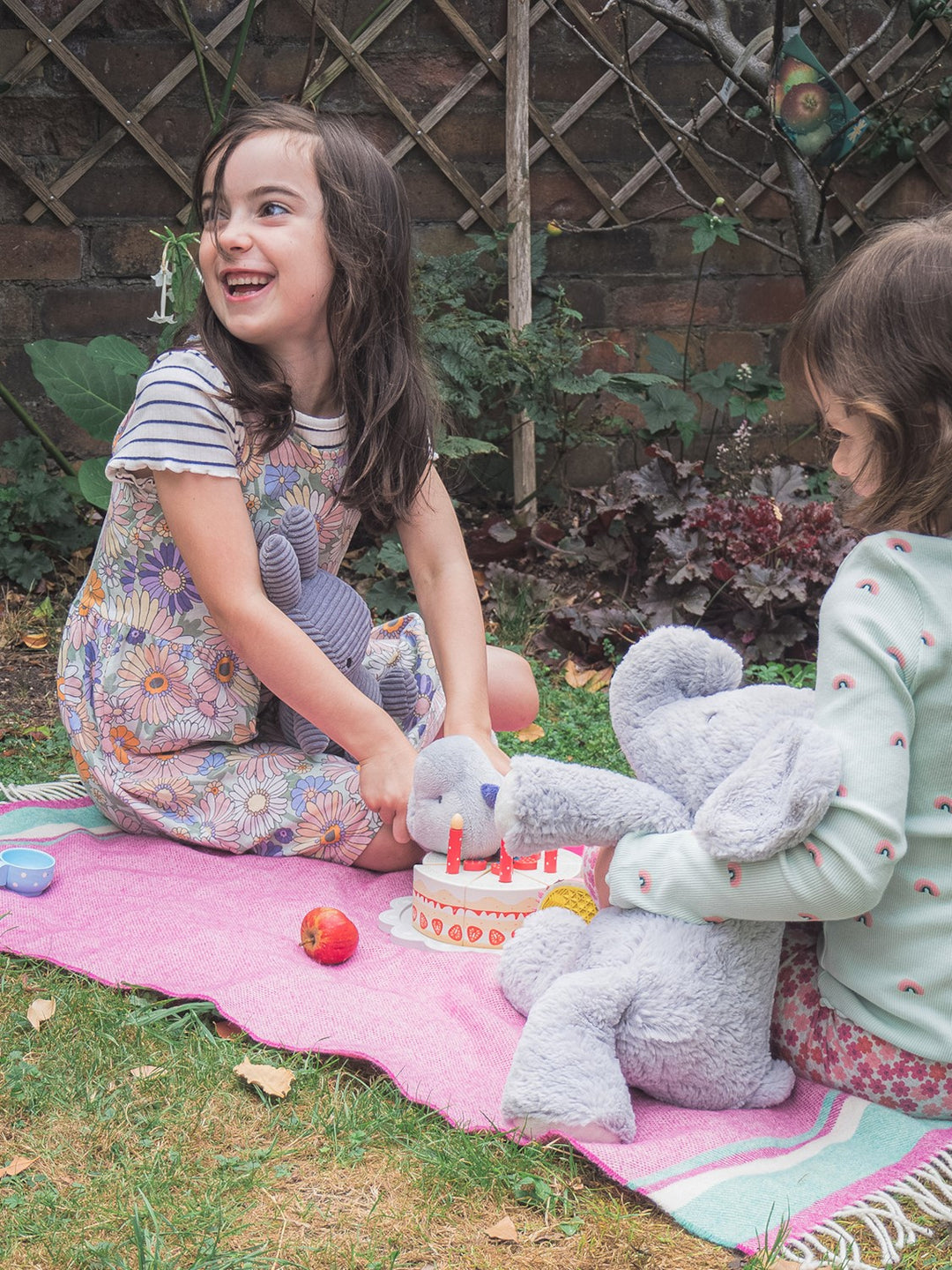 Two children in a garden sitting on top of a pink and blue merino lambswool baby blanket