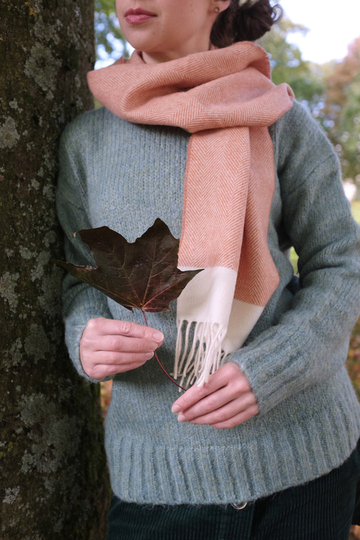A woman wearing an orange and cream wool scarf while leaning on a tree. 