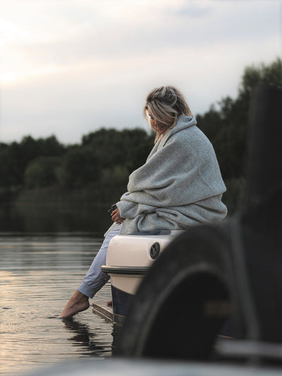 A woman wrapped with a large grey herringbone blanket dipping her feet in a body of water
