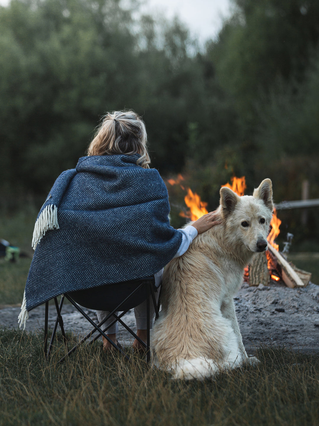 A woman wrapped with an extra large blue herringbone throw. She is sitting down facing a firepit next to a dog