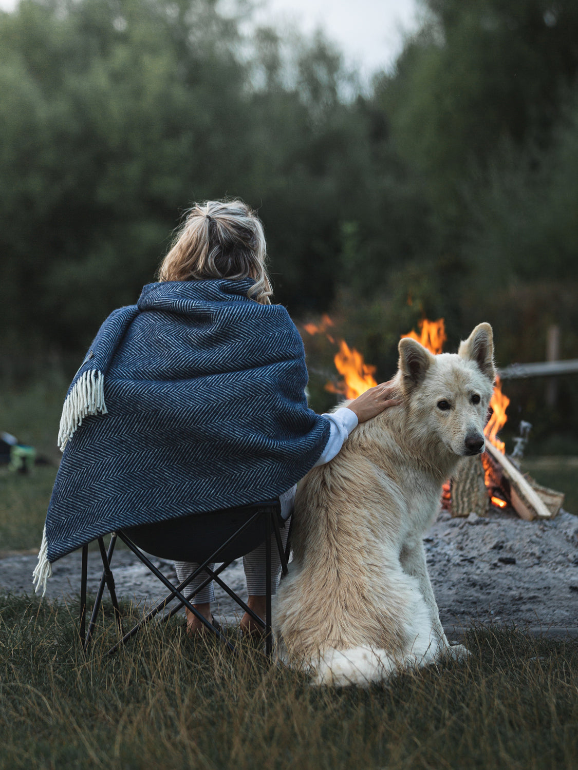 A woman sitting down on a chair facing a firepit wrapped in a navy blue wool blanket