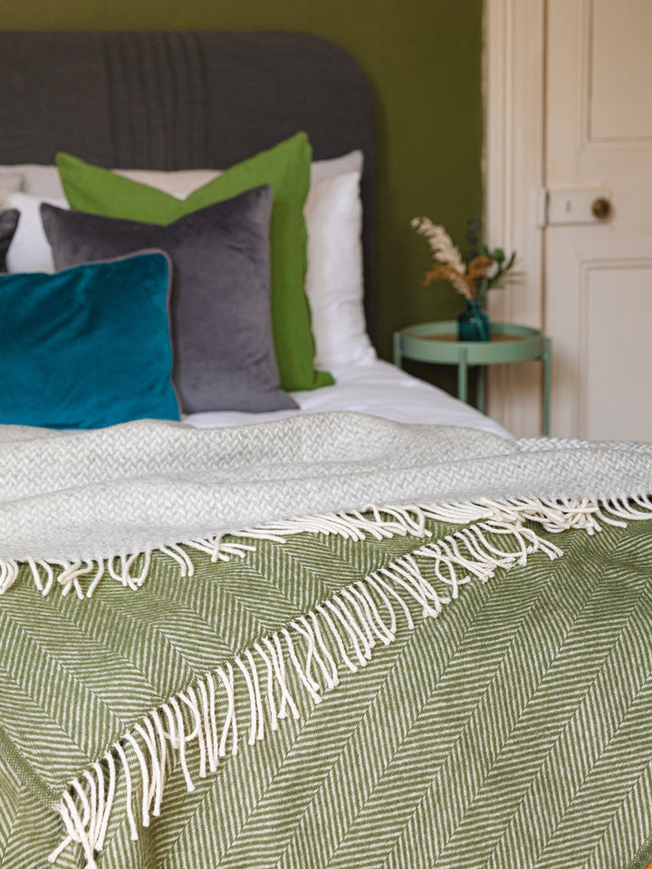Green herringbone wool throw draped across a bed with several cushions in the background