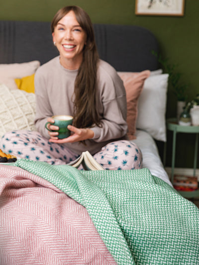 Green and pink wool blankets hanging off a bed. A woman sitting cross legged while holding a tea cup is in the background