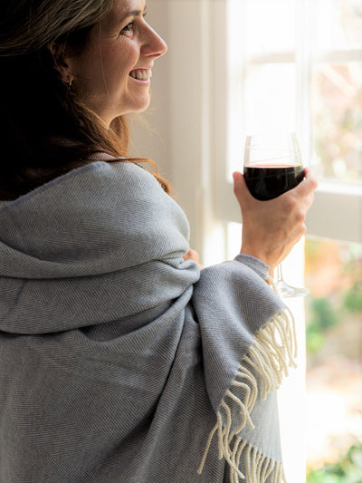 A woman wrapped with a large blue merino herringbone blanket holding a glass of wine