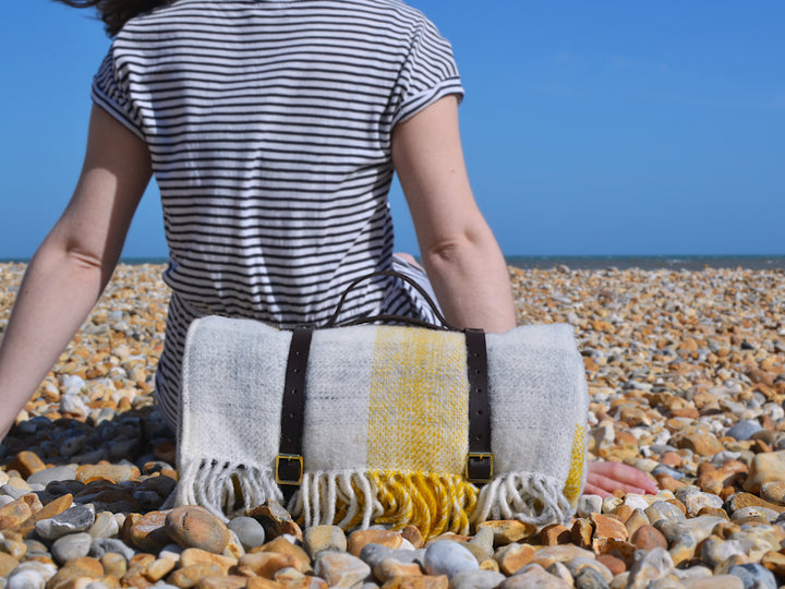 A rolled up yellow check wool picnic rug placed behind a person sitting down at a pebble beach