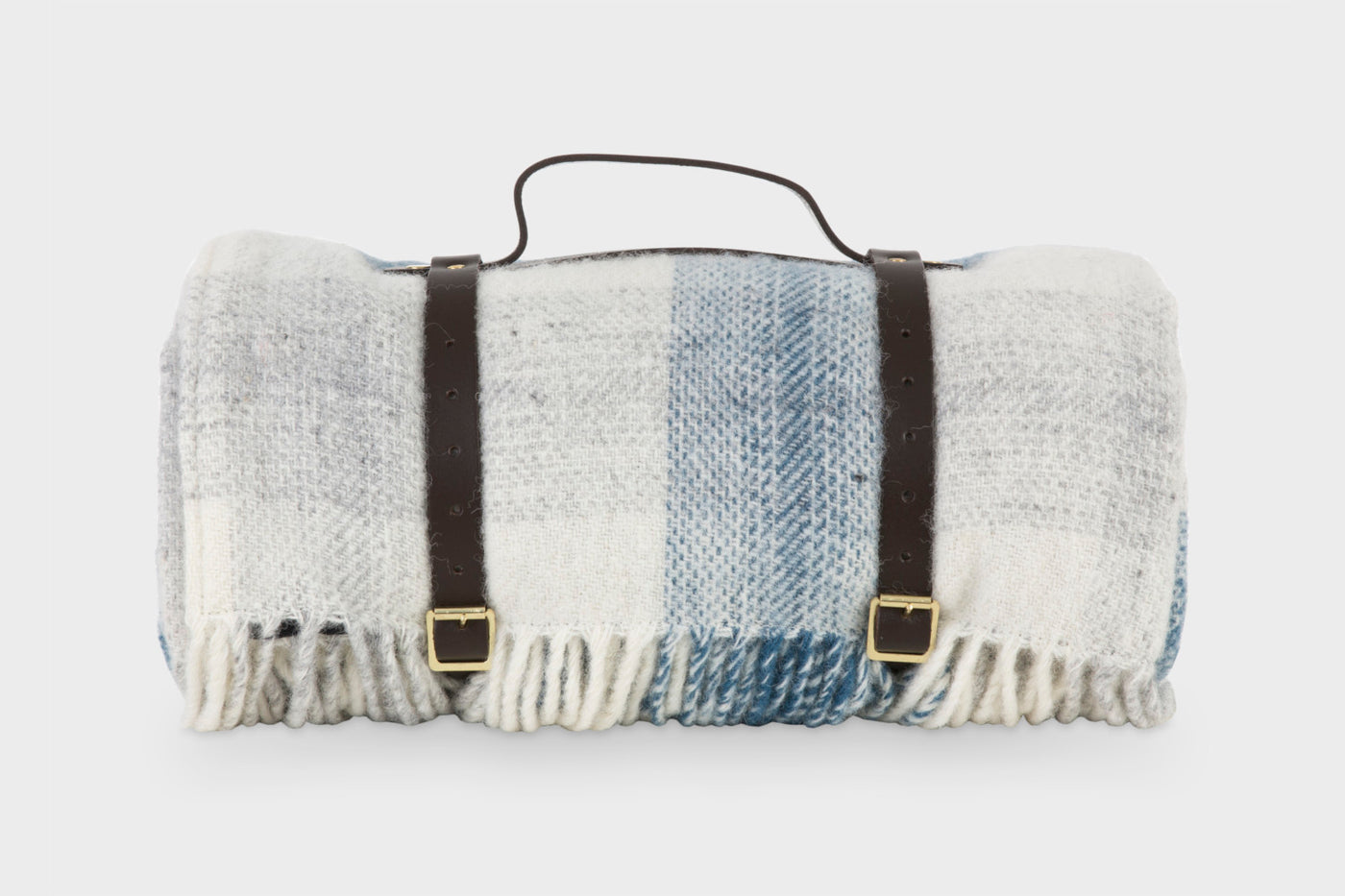 A blue and white wool picnic rug by The British Blanket Company. The picnic rug is rolled up with leather straps and a handle. 