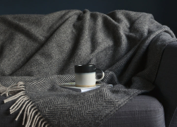 A mug and a book placed on top a grey herringbone wool blanket. The blanket is draped over a sofa. 