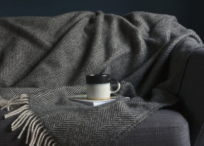 A mug and a book placed on top a grey herringbone wool blanket. The blanket is draped over a sofa. 