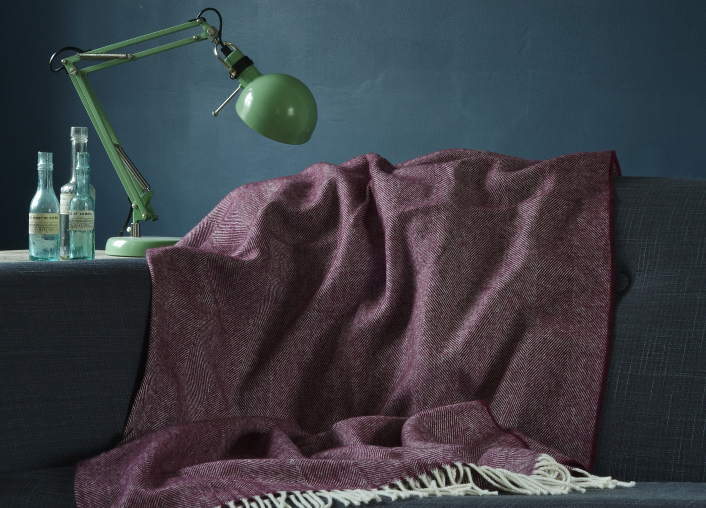 Large purple herringbone wool blanket draped over a sofa with green lamp and glass bottles on the left.
