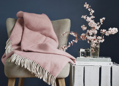 An extra large pink herringbone wool blanket draped over a lounge chair on the left. A glass filled with pink flowers, a camera, and books are placed on a wooden crate on the right