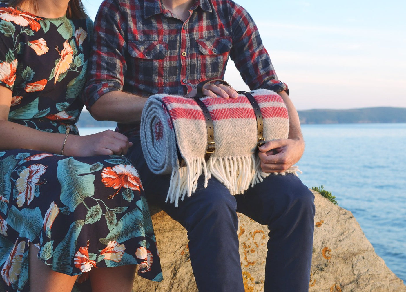 A man and woman sitting side by side while the man holds a rolled up wool picnic rug.