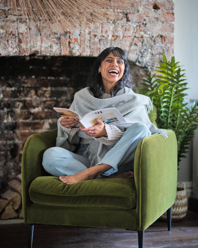 laughing woman sitting on a green armchair wearing a light grey wearable blanket wrap made from pure wool by The British Blanket Company online shop