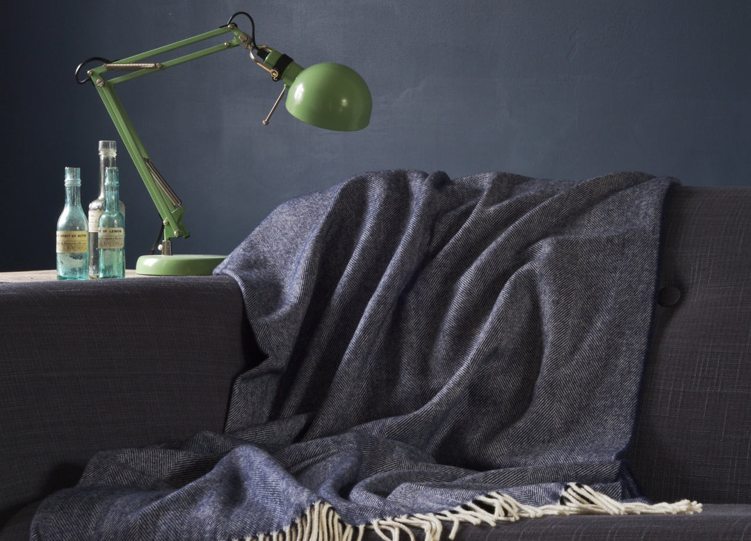 Large blue merino herringbone wool blanket draped over a sofa. A table lamp and glass bottles are on top a table next to the sofa