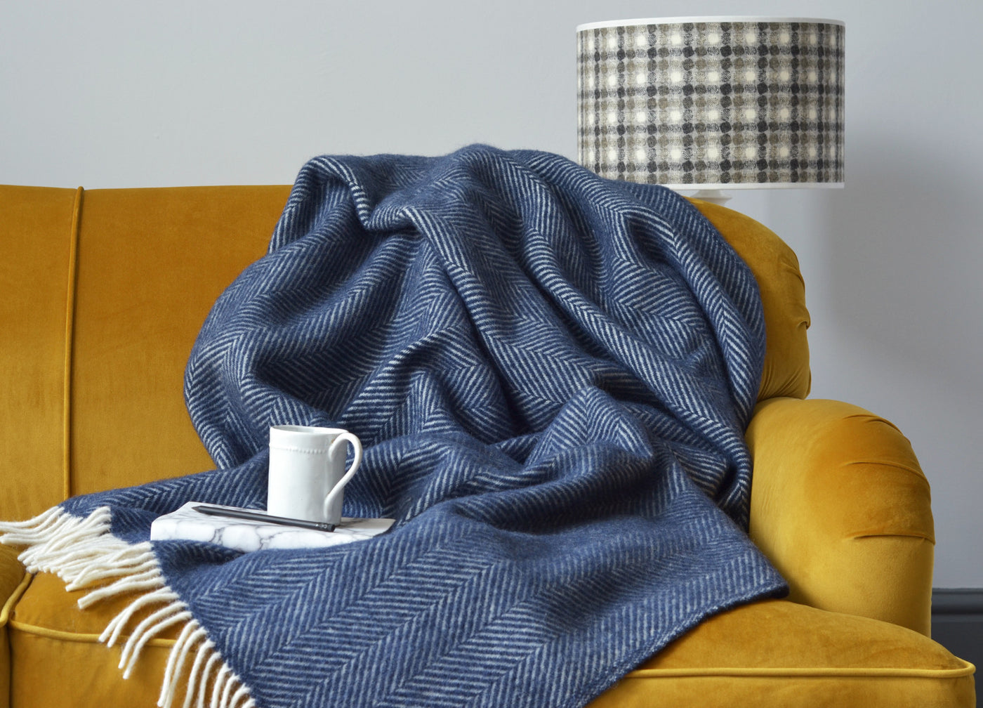 Large navy blue herringbone wool blanket draped over a yellow sofa. A mug and book are placed on top of the blanket