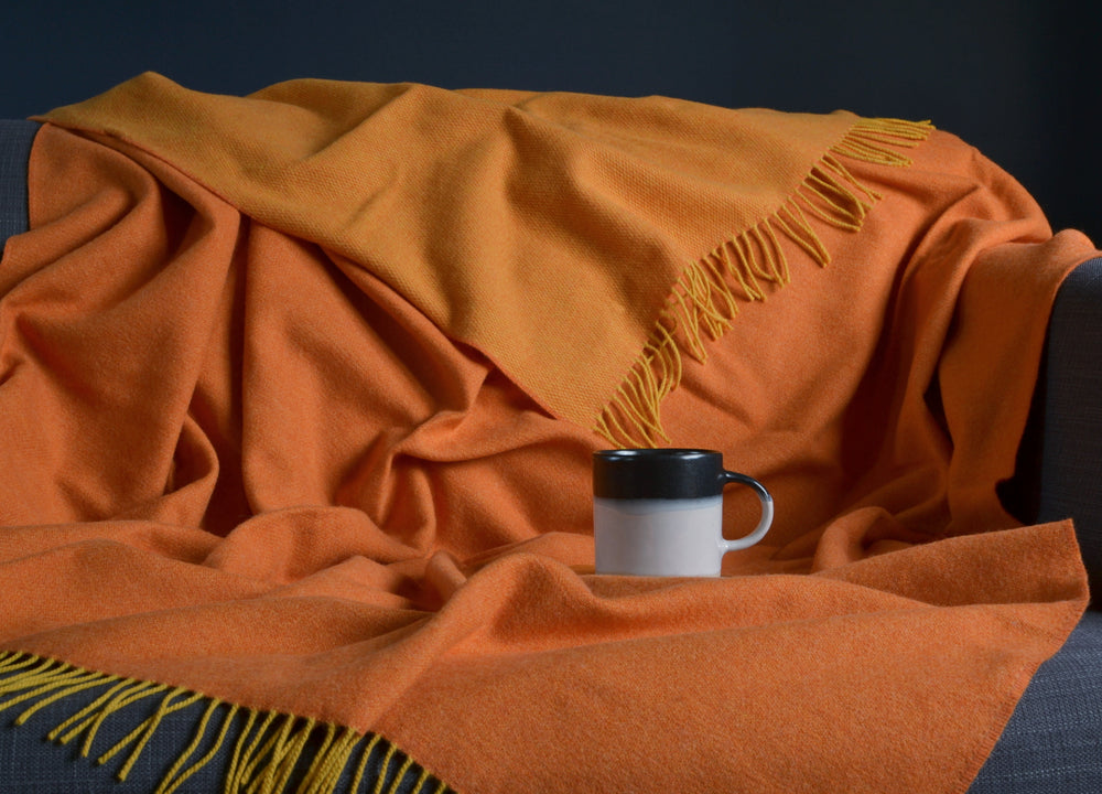 Large orange and yellow reversible merino wool blanket draped over a sofa with a mug on top the blanket