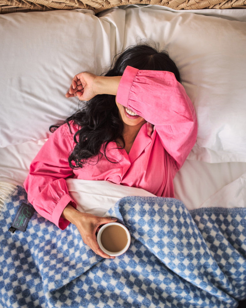 smiling woman wearing pink pyjamas lying in bed and covered in blue spot wool throw blanket by The British Blanket Company online shop