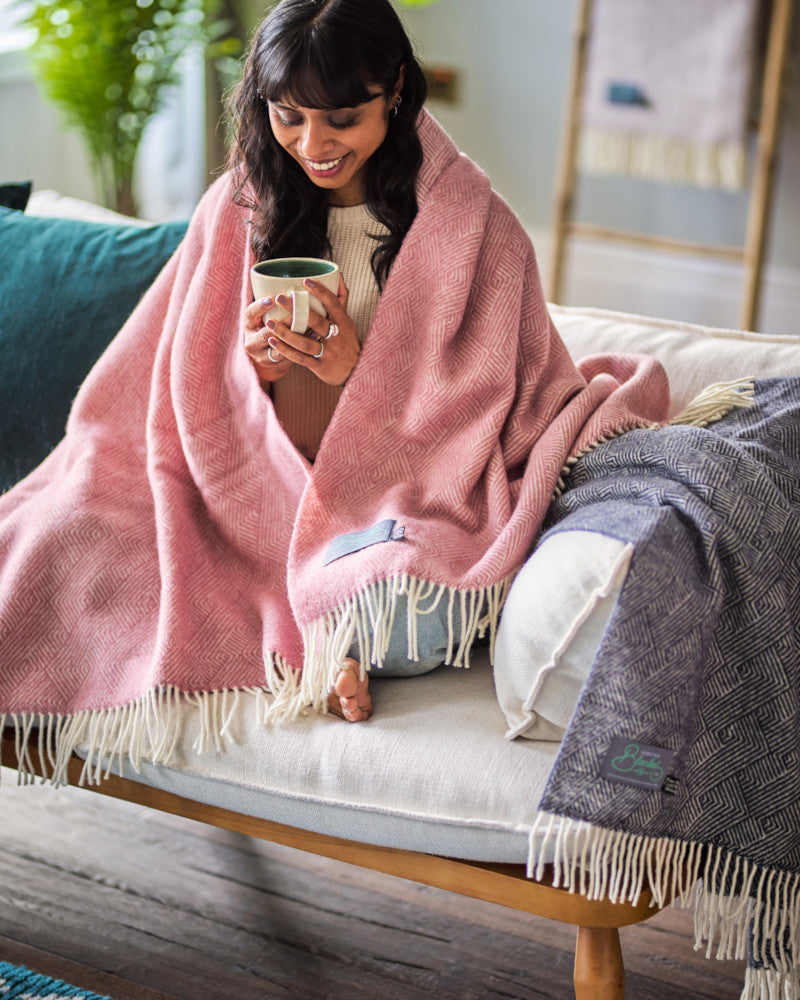 smiling woman drinking tea on sofa wrapped in pink pure wool throw blanket by The British Blanket Company online shop