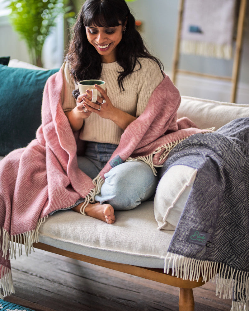 smiling woman drinking tea on a sofa wrapped in pure wool throw blankets by The British Blanket Company online shop