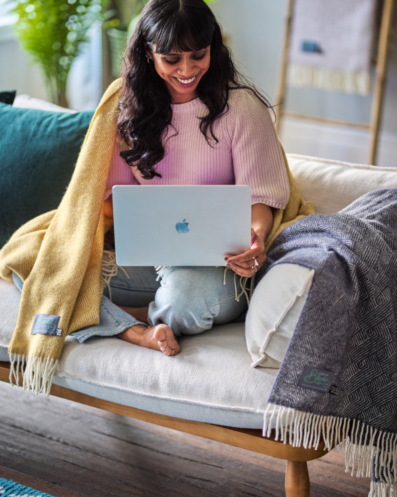 smiling woman working on laptop on a sofa wrapped in a yellow wool throw blanket by The British Blanket Company online shop