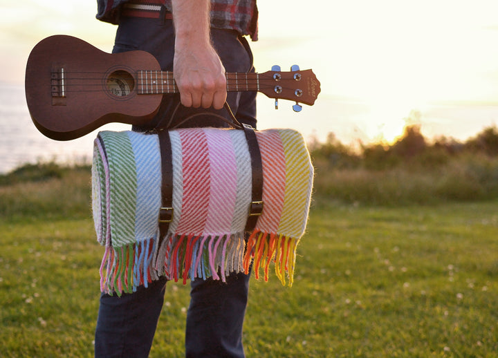 A man holding a ukulele and the handle for leather straps carrying a rainbow stripe wool picnic rug