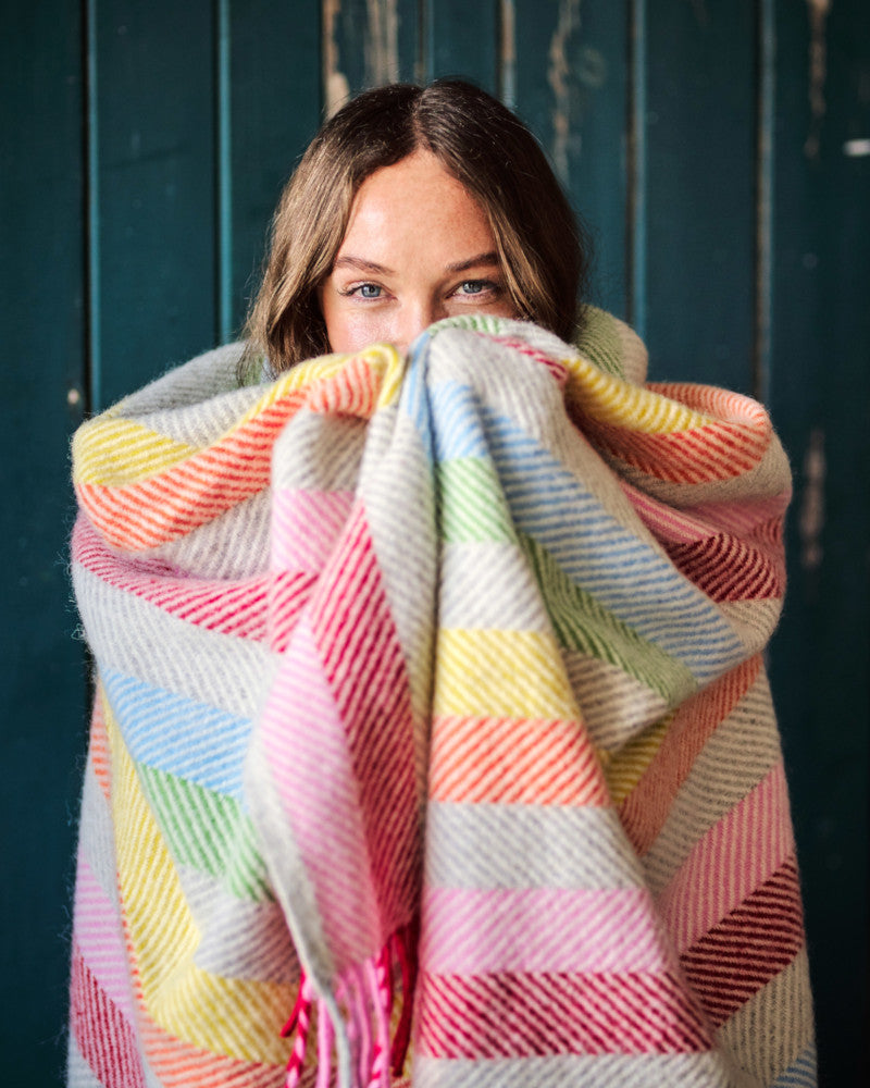 smiling woman wrapped up in rainbow stripe pure wool herringbone throw blanket by The British Blanket Company online shop