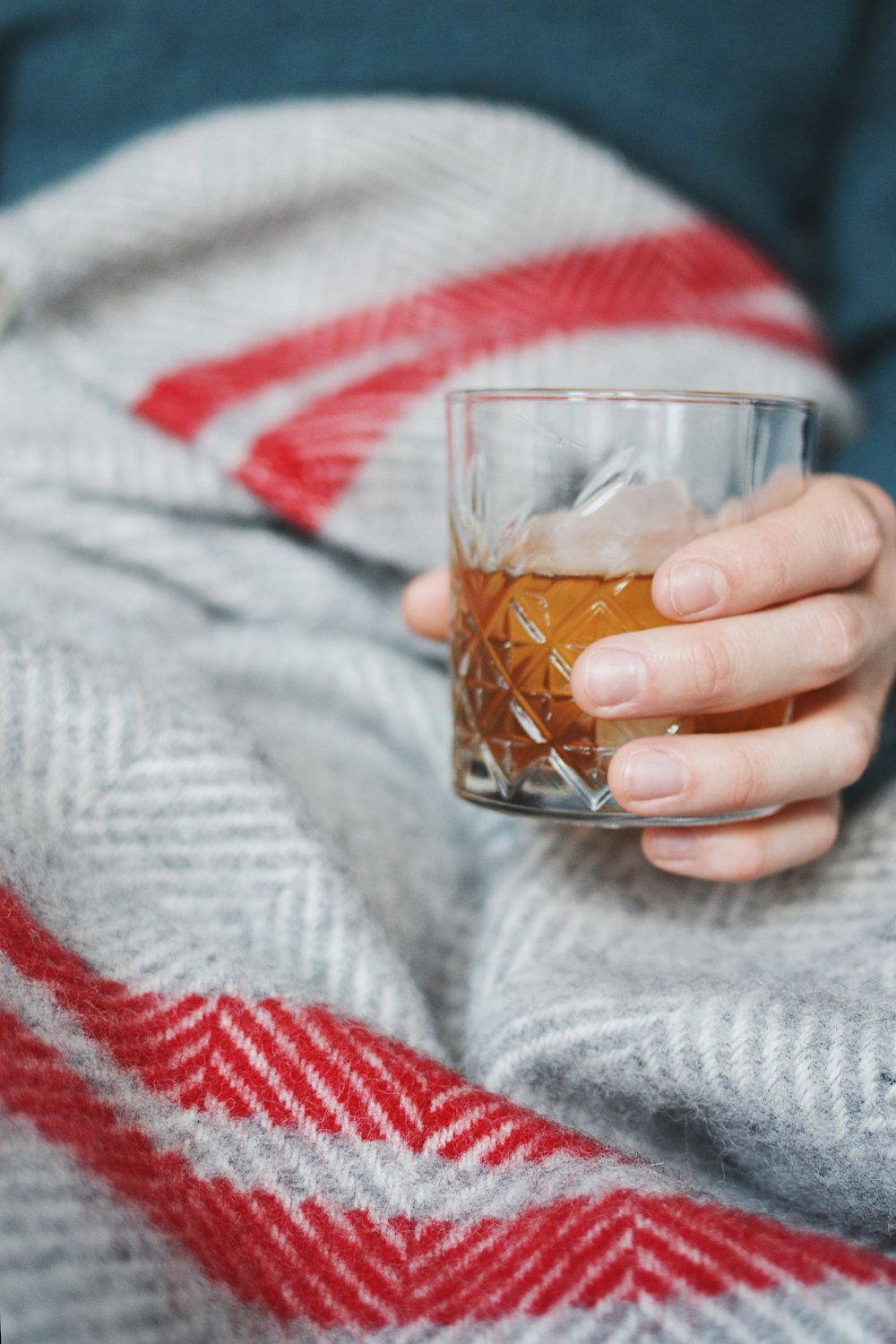 A person holding a glass with a red and grey wool picnic blanket draped over them.