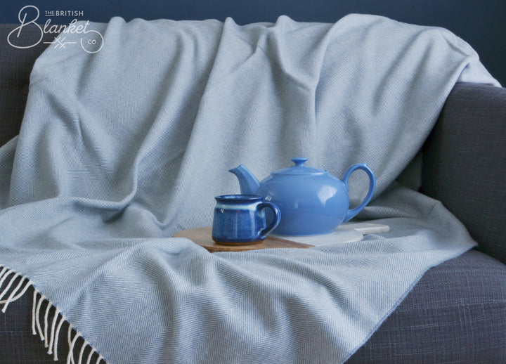 A large blue merino herringbone wool blanket draped over a sofa. A mug and tea pot are on top of the blanket