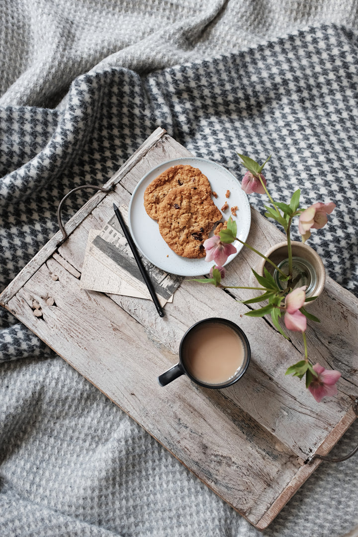Wooden tray carrying a plate of biscuits, mug, and flowers is placed on top of wool blankets in various shades of grey