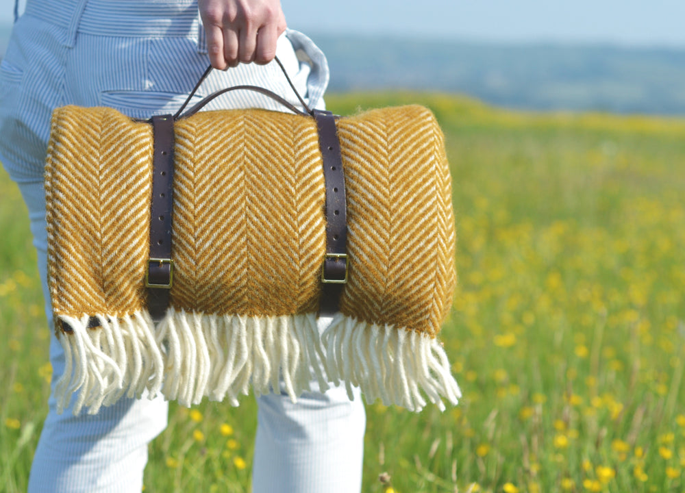A person holding the handle of leather straps carrying a yellow herringbone wool picnic rug